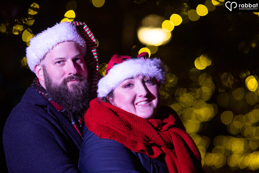 Couple looking at camera in front of lit up Christmas tree.
