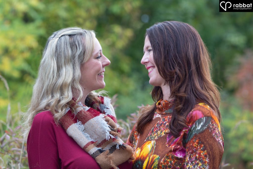 Lesbian couple looking each other in the eyes and smiling lovingly. Greenery in background. Close-up portrait.