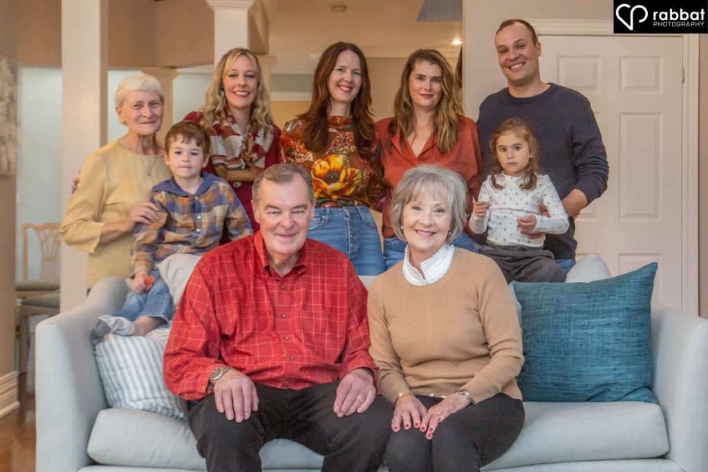 Matriarch and patriarch on light couch with ocean blue pillow and their family behind them including two young children, the matriarch's sister, their two children and their partners.