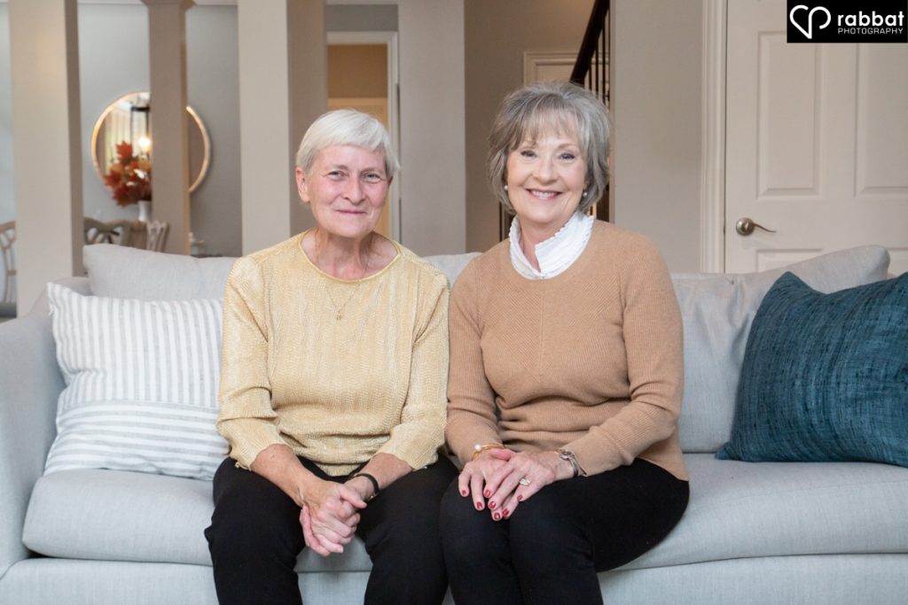 Two sisters in their 60s or 70s sitting on a couch beside each other.