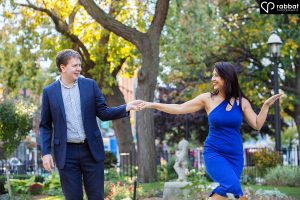 Blonde man with South Asian Woman in a dance pose in front of trees. He is wearing a suit, she is wearing a short sleeve blue dress.