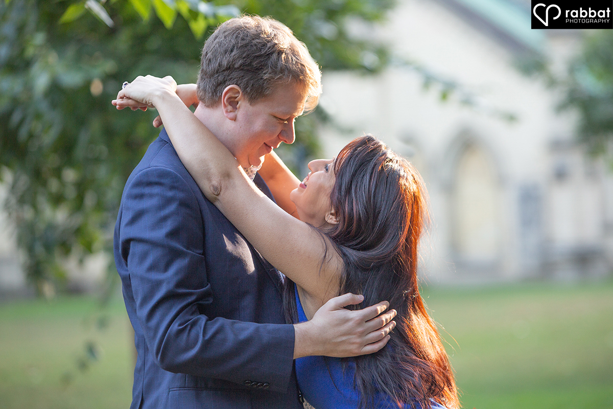 Light haired man and South Asian Woman. Her arms are on his shoulders and her hands are clasped at the top. His hands are around her shoulders, as if he is about to lead her into a dance. He is wearing a suit and she is wearing a dark blue dress. There is a tree and out of focus church behind them.