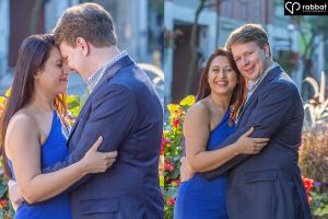 Engaged couple holding each other at the waist. He is blonde, she is south Asian. she has long brown hair. They are looking straight at each other. She is wearing a blue dress. He is wearing a suit. In the second photo, they are both looking at the camera. They have red flowers behind them as well as an out of focus Toronto store.