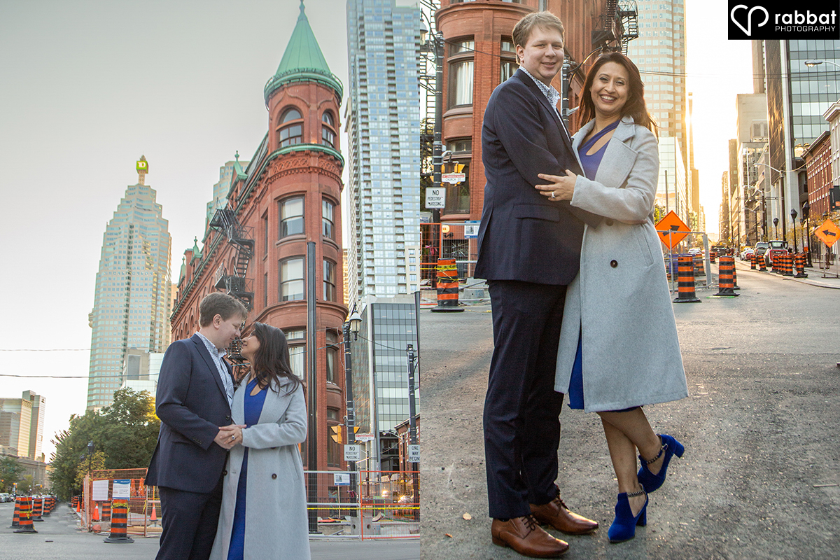 South Asian woman in long gray coat with blue high heel shoes and white man hugging in front of flat iron building in downtown Toronto.