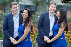 Two vertical photos side by side of a white man in a suit with an open jacket holding hands at the bottom with a South Asian woman in a blue dress. In the first photo, they are both looking at the camera. He is smiling and she is laughing. In the second photo, he is smiling and looking straight ahead at the camera and she is looking at him. Photos are in front of a flower bed with downtown Toronto stores behind them.