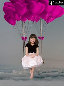Photo of a 6 year old in a dress with a black velvet T-shirt top and pink silky skirt. She is sitting on a swing above the clouds with heart balloons above her head.