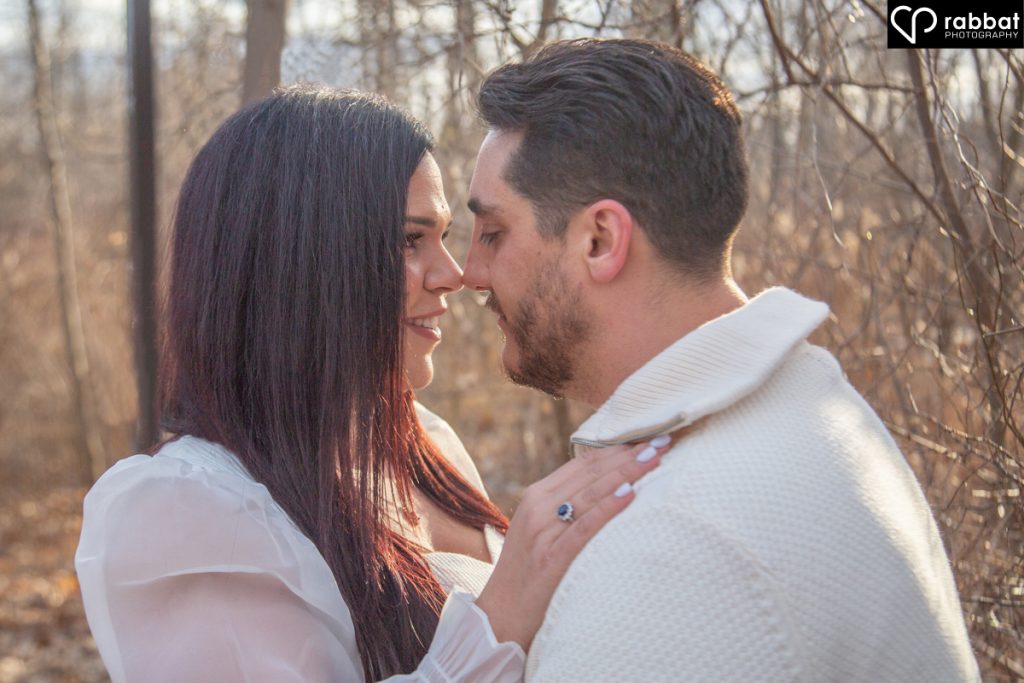  Man and woman holding hands both wearing white shirts with rim lighting in background. They are head to head and you can only see from the tops of their heads, to their elbows. Her engagement ring is showing. Forest background