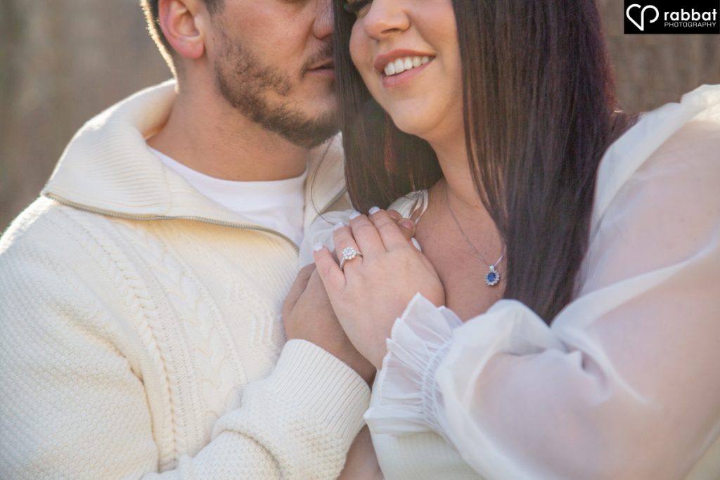 Man and woman holding hands both wearing white shirts with rim lighting in background. They are head to head and you can only see from the tops of their heads, to their elbows. Her engagement ring is showing. Forest background