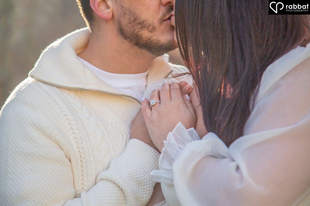 Man placing lips and nose behind woman's hair. Only their noses to elbows is shown. They are both wearing white. Her hand is on top of his. Forest background.