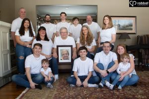 16 person family portrait, 17 including framed photo of deceased wife/grandma. They are all wearing white t-shirts.