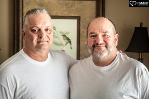 Two grown men in white t-shirts smiling side by side.