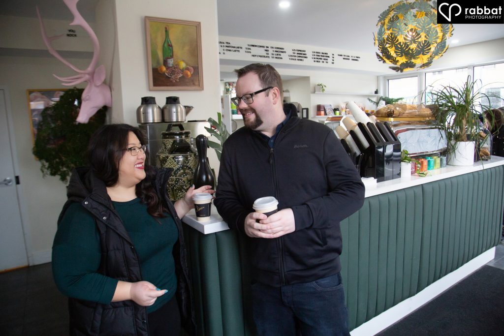Couple looking at each other while holding coffees inside a local coffee shop. Asian woman, white man. THey are smiling at each other. The store has a pink moose head on the wall behind them and the counter is green. The couple is in front of the counter.