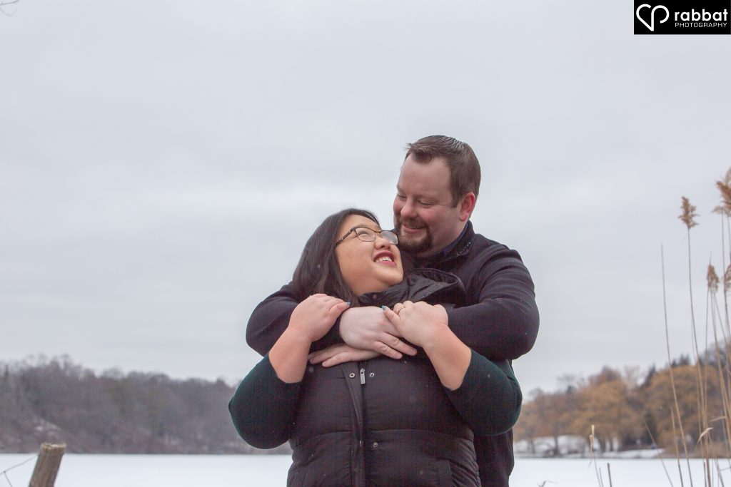 White man hugging Asian woman from behind, in front of Grenadier pond. SHe is looking behind her to smile at him and he is looking at her.