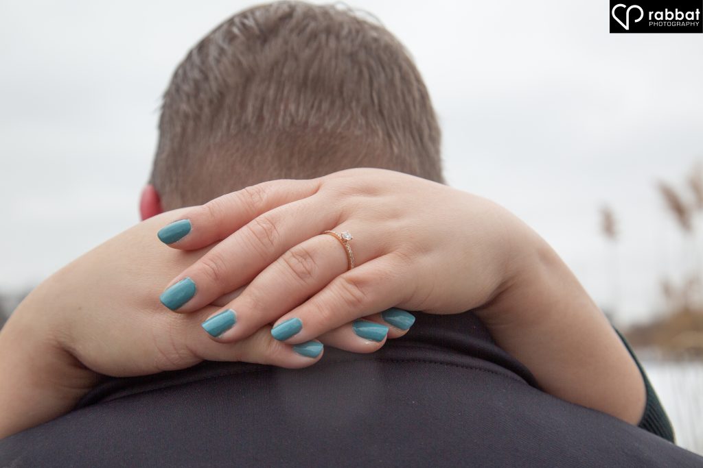 Woman's hands wrapped around the back of a man's head. She has turquoise fingernails and you can see her diamond wedding ring. It is a single diamond on a rose gold band.