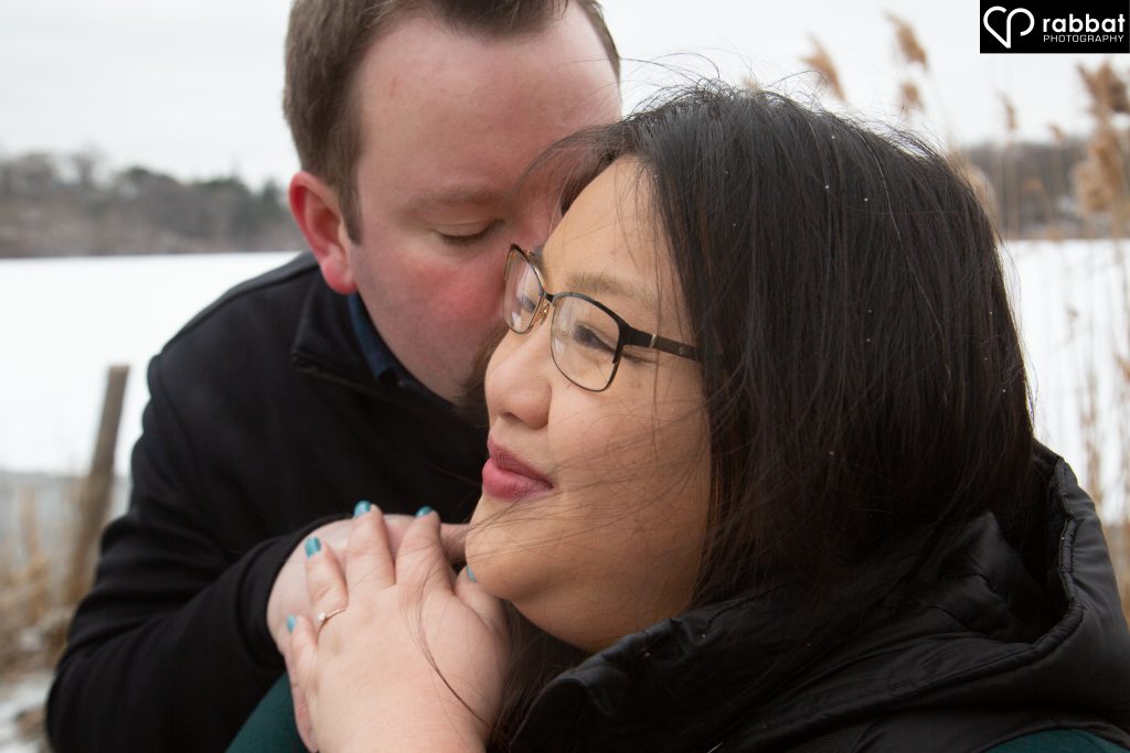 Close up of white man kissing Asian woman in front of Grenadier pond. They are both wearing black. You can see the pond and the out of focus trees in the background.