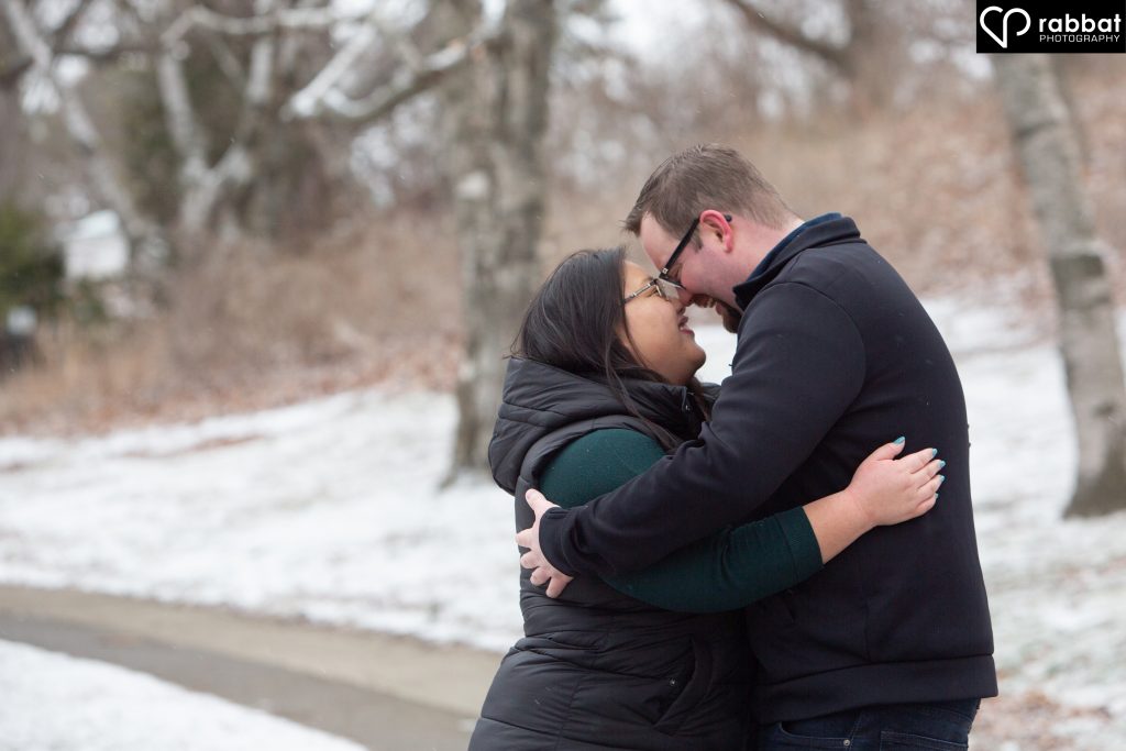Asian woman and white man with foreheads touching as they hug each other on a snowy path with barren trees.