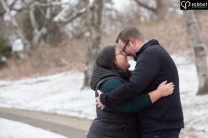Asian woman and white man with foreheads touching as they hug each other on a snowy path with barren trees.