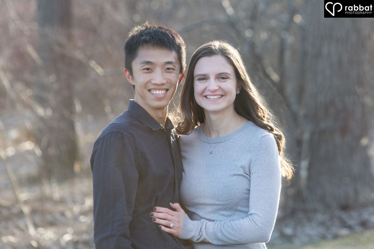 Couple looking at camera with forest behind them. Man is Asian, woman is white. He is wearing a dark shirt and she is wearing a bluish gray long sleeve shirt. They are smiling at the camera and are backlit by the sun.