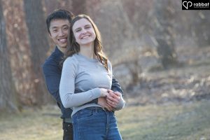 Couple looking at camera with forest behind them. Man is Asian, woman is white. He is wearing a dark shirt and she is wearing a bluish gray long sleeve shirt. They are smiling at the camera and are backlit by the sun. Man is behind woman, hugging her at the waist. Her hands are clasped around her waist on top of his.