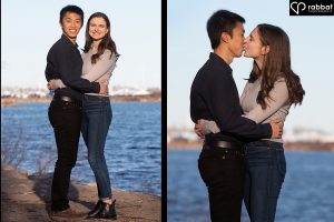 Engagement photos at Cherry Beach. Side by side vertical photos of a couple standing in front of Lake Ontario on a sunny evening with a blue sky in the background. In the photo on the left, you can see their full bodies and they are hugging and looking at the camera. In the photo on the right, they are kissing and they are shown from just above the knees. Man is Asian, woman is white with brown hair. They are both wearing black pants. He is wearing a long-sleeve black shirt and she is wearing a long-sleeve grayish blue shirt.