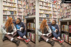 Two side by side vertical photos of a couple sitting on floor in front of bookshelves filled with comics and anime. They are each holding a comic book. The books are open and they are looking at each other in both photos. Woman is wearing a jean jacket, short skirt and black boots with white socks. Man is wearing a jean jacket. Man is Black and woman is Metis with a light complexion and red hair.