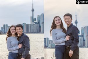 Cherry Beach engagement session. Vertical photos of a couple with the Toronto skyline including Lake Ontario and the CN Tower in the background. They are smiling at the camera. The woman is wearing a long sleeve grayish blue shirt and the man is wearing a dark shirt. The man is Asian and the woman is white with brown hair.