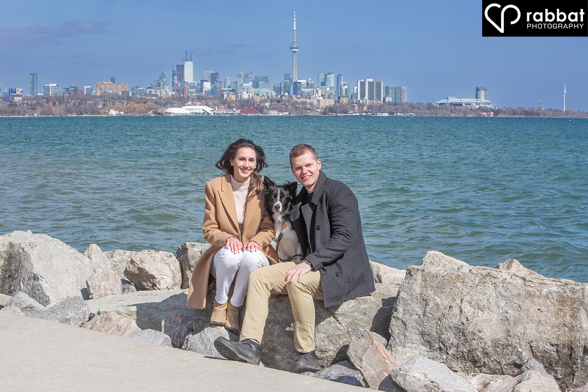 Couple in front of Lake Ontario sitting on rocks with their dog licking the woman's face and the Toronto skyline in the background. The water is very blue.