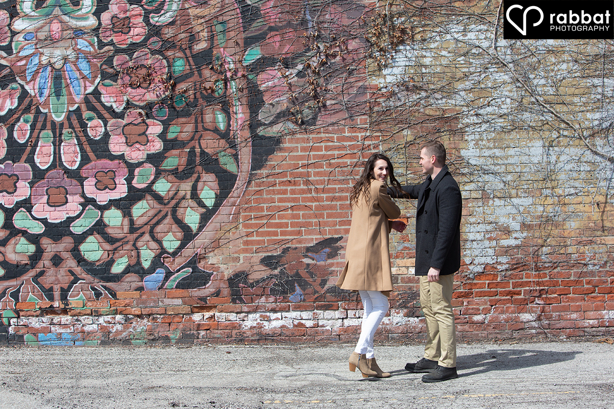A woman with a brown coat, white pants and brown suede boots about to be twirled by a man in a black coat and beige pants in front of a beautiful, colourful floral mosaic mural on a red brick wall.