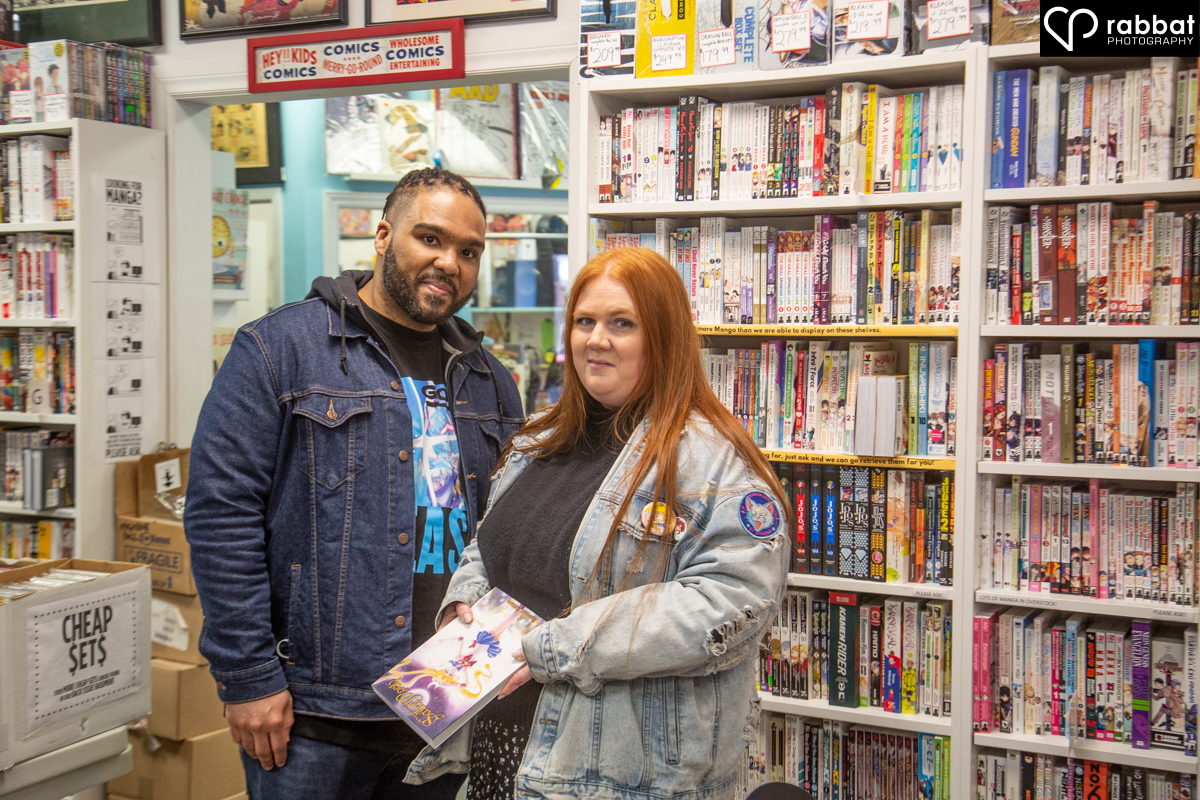 Couple holding a comic book in front of shelves full of books. They are looking at the camera. Man is black and woman is Metis with a light complexion and red hair. They are both wearing jean jackets/.