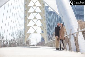 Wide angle photo of man in black coat with beige pants and woman in beige coat with white pants with a dog in front of them on the Humber Bay bridge. They are looking at the camera and standing on the right of the bridge.