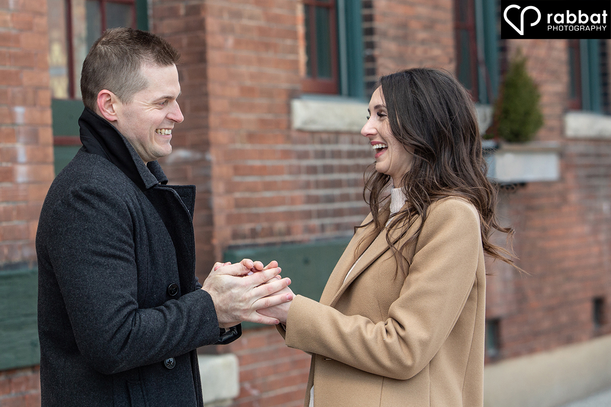 Man with short brown hair warming up the hands of woman with curly long brown hair. He is on the left and she is on the right and they are facing each other and close to laughter. They are against a red brick wall with windows that have green around them.