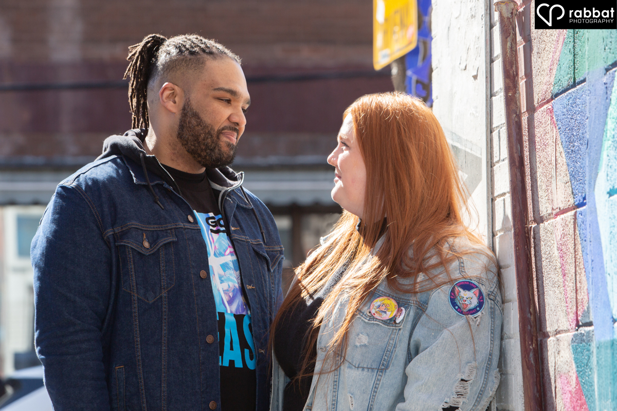 Backlit couple in front of an artistic mural looking lovingly at each other. Man is black and woman Metis but has a light complexion with red hair. Woman is wearing a jean jacket. Man is wearing a jean jacket with a T-shirt underneath.