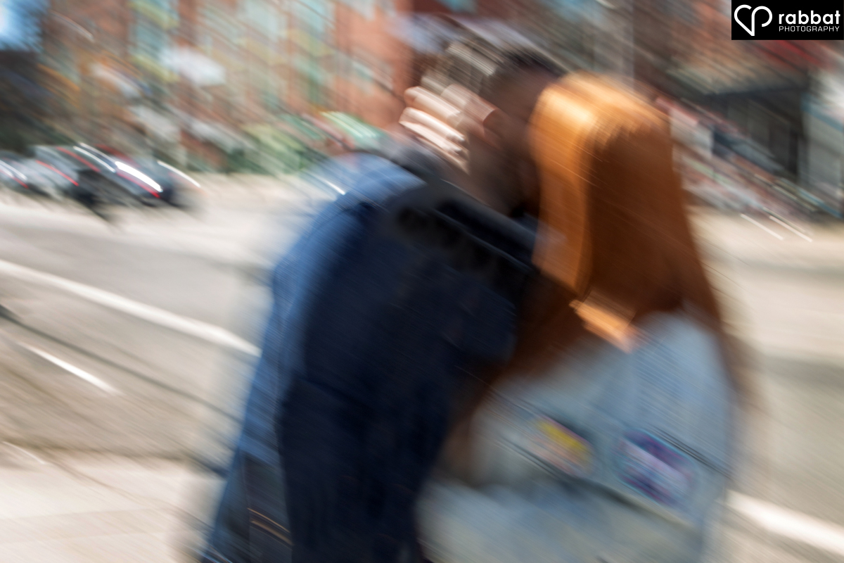 Blurred photo of a couple in jean jackets kissing in front of a road. Her long red hair stands out. This is an impressionist style photo.