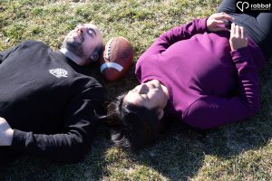 Sporty engagement photo. White man in black long sleeve shirt lying beside South Asian woman in purpleish pink shirt with football in-between them. Their eyes are closed and the photo was taken from above.