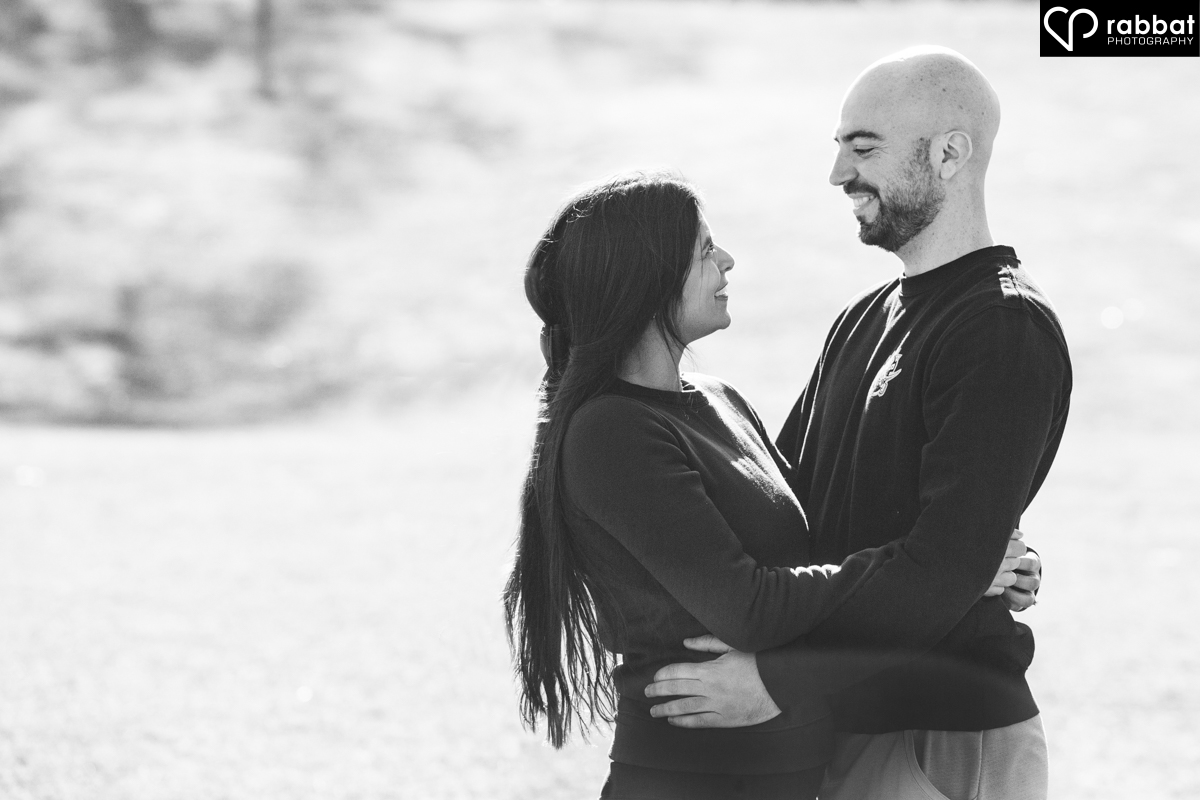 Black and white photo of a couple looking into each other's eyes and smiling. They are surrounded by a grassy field and it is a sunny day.