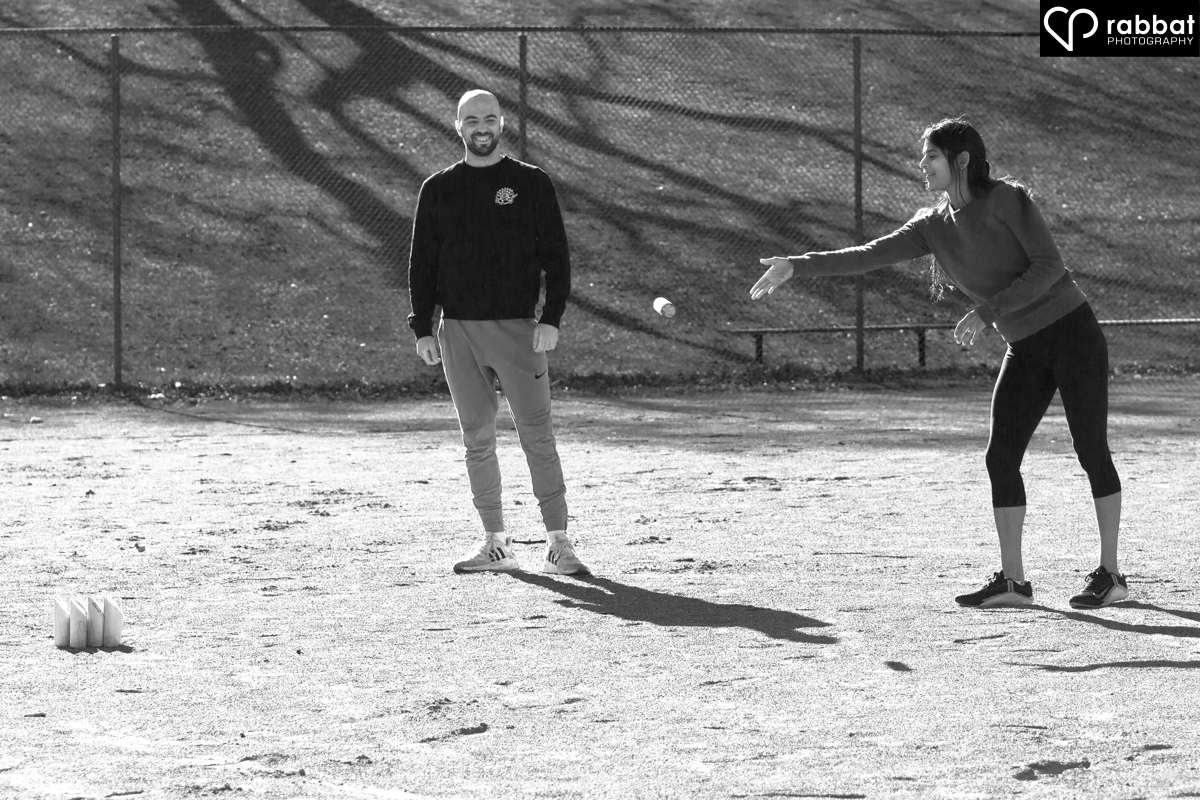 South Asian woman throwing a Molkki stick in a baseball field as her white fiance looks on and smiles. Black and white photo in baseball diamond.