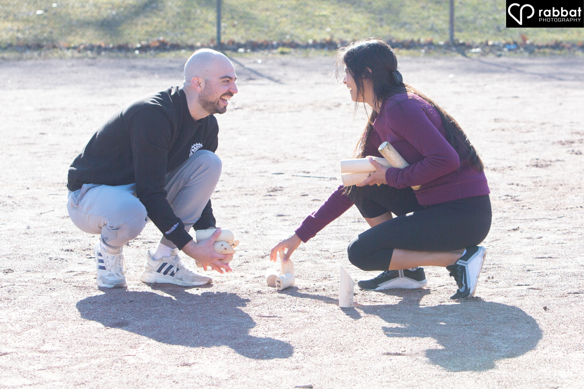 Colour photo of engaged couple picking up Molkky sticks in a baseball diamond.