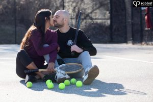 Sporty engagement photo. Couple sitting on tennis court. They are each holding wooden tennis courts. They have lots of tennis balls in front of them. They are looking at each other and smiling. Woman is South Asian, wearing a purple shirt and black leggings. Man is white, wearing a black long sleeved shirt and blue jeans. They are kissing.