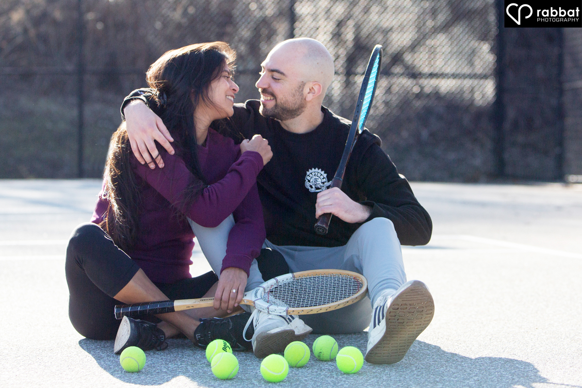 Couple sitting on tennis court. They are each holding wooden tennis courts. They have lots of tennis balls in front of them. They are looking at each other and smiling. Woman is South Asian, wearing a purple shirt and black leggings. Man is white, wearing a black long sleeved shirt and blue jeans.