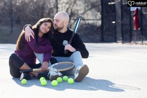 Sporty engagement photo at Jeff Healey Park. White man hugging South Asian woman on tennis court. They are each holding wooden tennis courts. They have lots of tennis balls in front of them.
