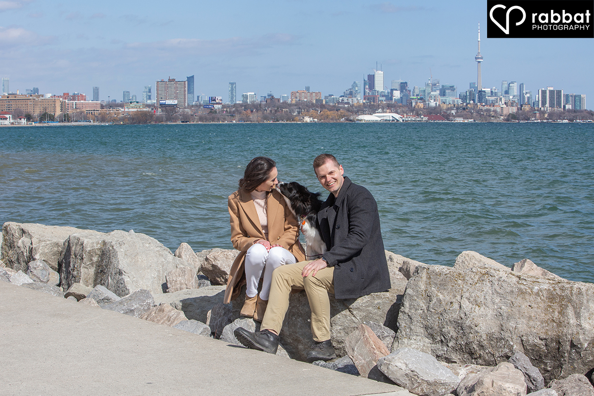 Couple in front of Lake Ontario sitting on rocks with their dog licking the woman's face and the Toronto skyline in the background. The water is very blue.