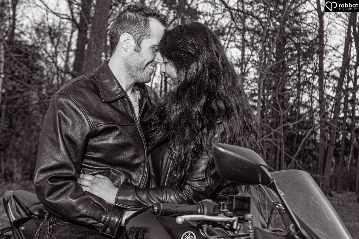 Black and white photo of couple on motorcycle with woman facing backwards. Their foreheads are touching. 