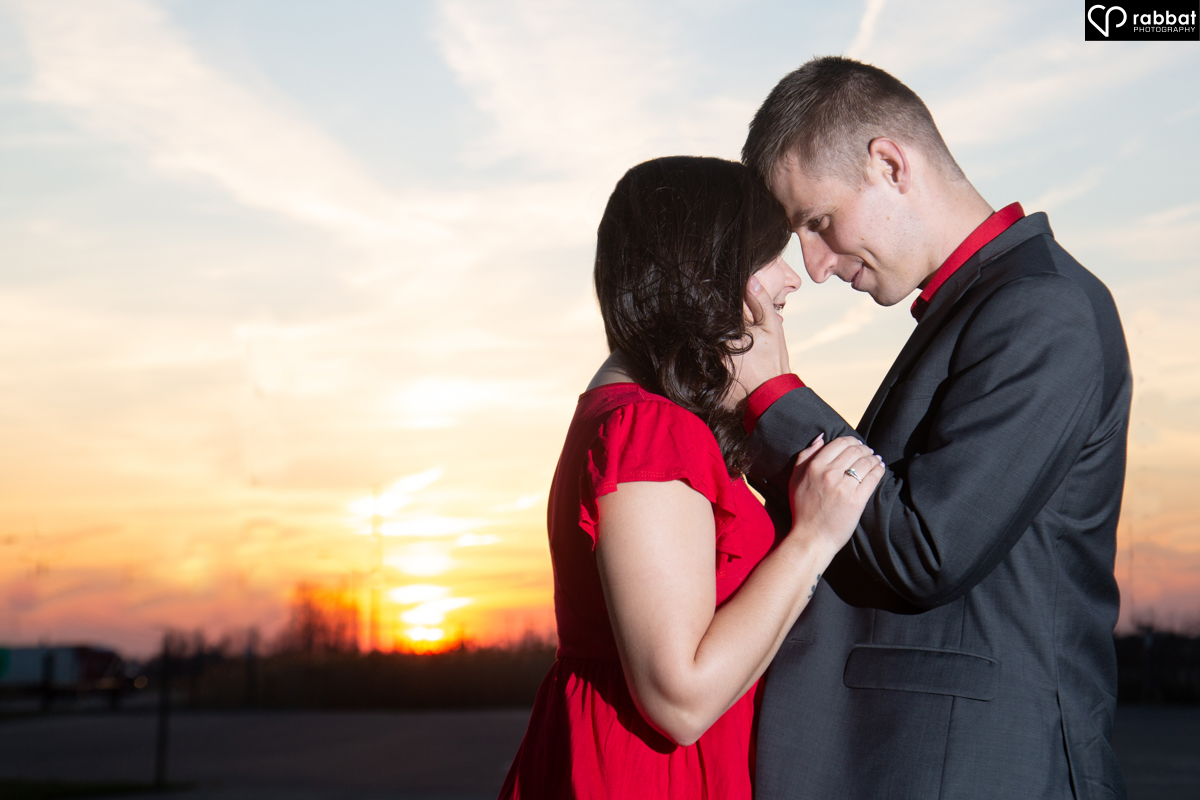 Woman in red dress and man in suit in front of sunset. He is holding her head, she is holding his arms and they are forehead to forehead.