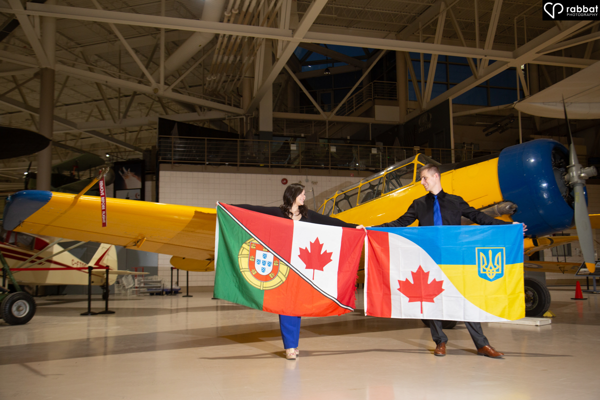 Couple holding up Portuguese and Canadian as well as Ukrainian and Canadian flags in front of yellow Royal Canadian Airforces airplane, indoors.