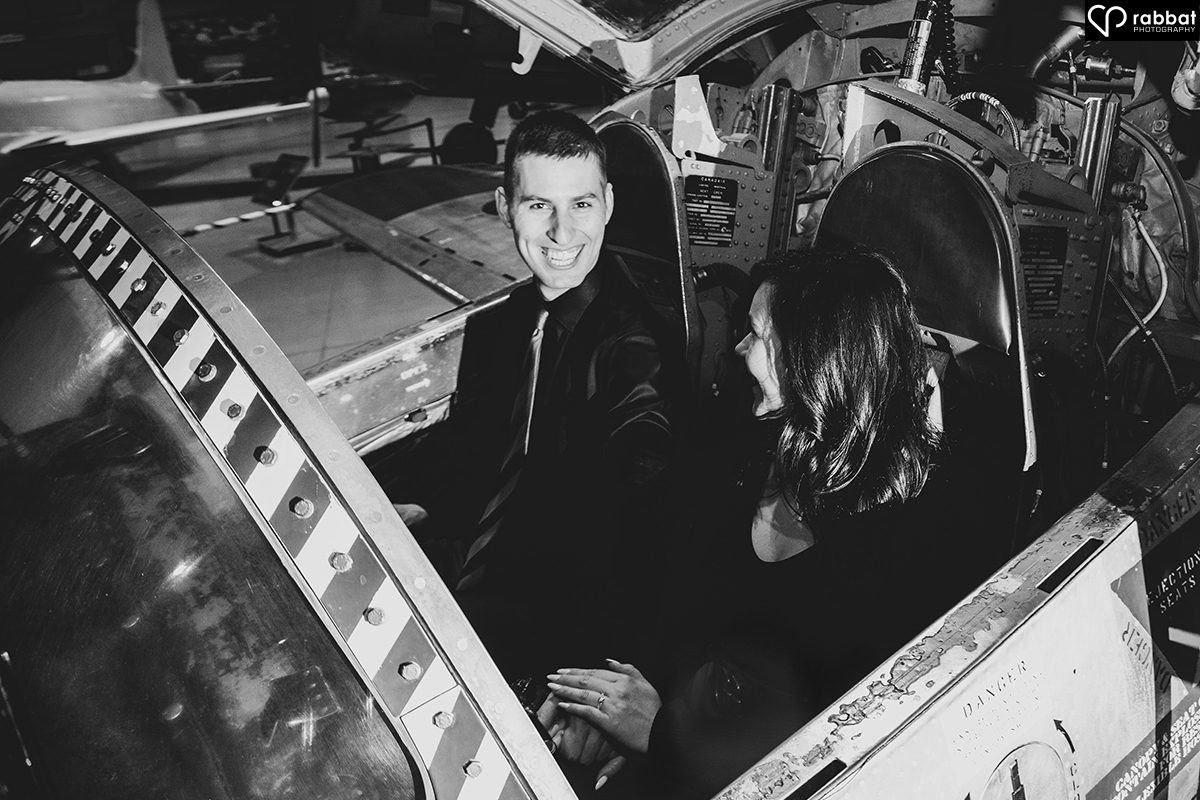 Black and white photo of a couple inside an airplane. The woman is looking at the man and the man is grinning a bit at her, and a bit at the camera.