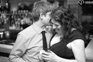 Black and white photo of a man whispering something in smiling woman's ear in front of a bar. She is holding a drink with a lemon slice.