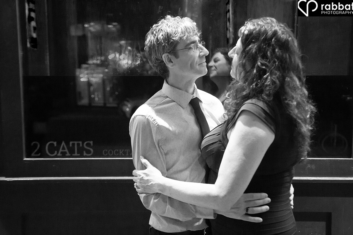 Black and white photo of a couple looking lovingly at each other in front of Two Cats Bar.