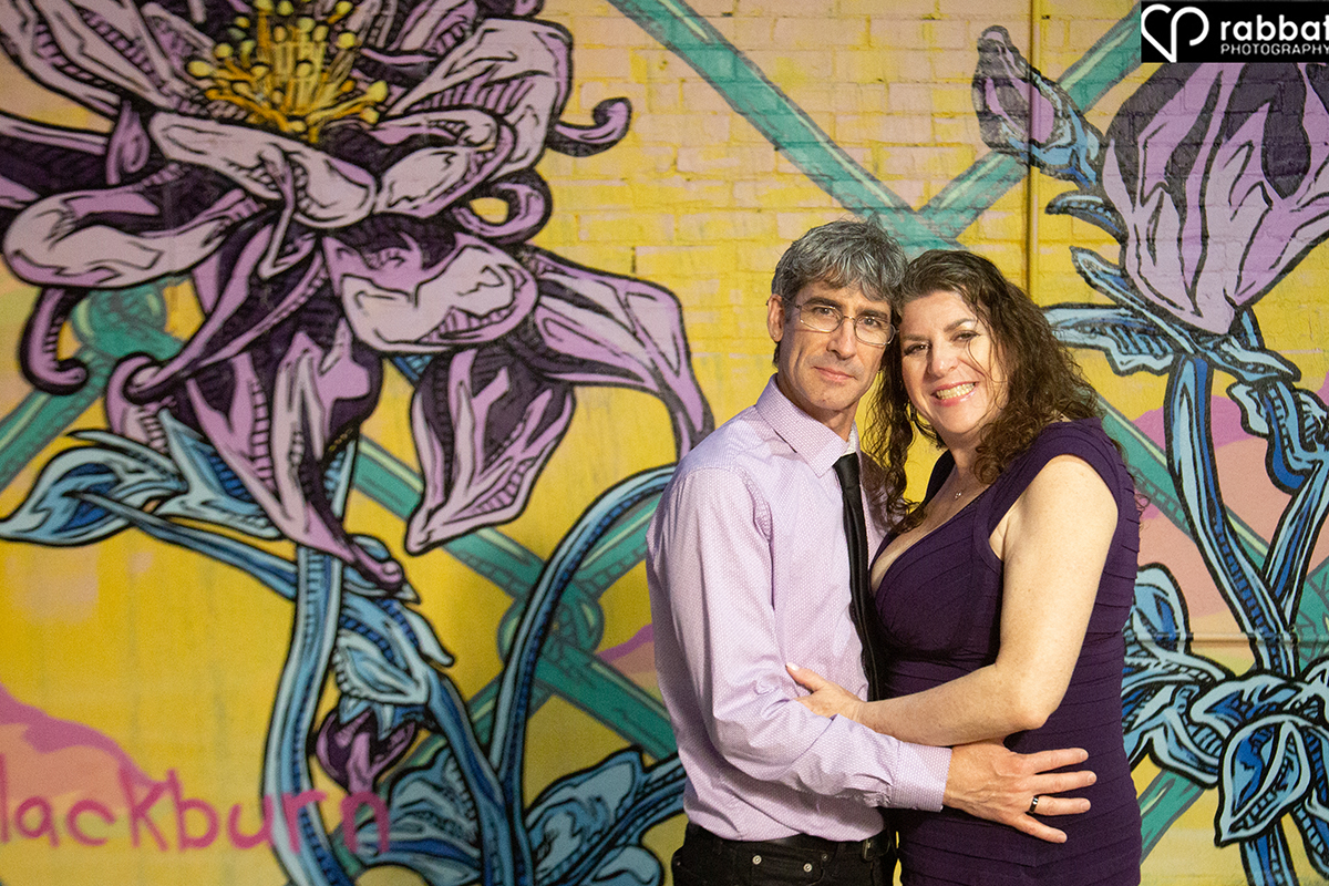 Couple holding each other at waist and looking at camera in front of a mural of giant Iris flowers. They are both wearing purple.